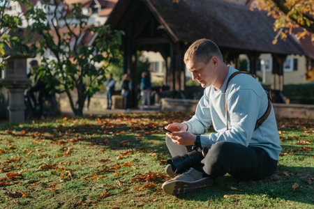 Professional Photographer Taking Picture Of Beautiful Autumn Park. Man Professional Photographer Sit With Camera And With Smartphone In Autumn Park. Retouched, Vibrant Colors, Brownish Tones.の写真素材