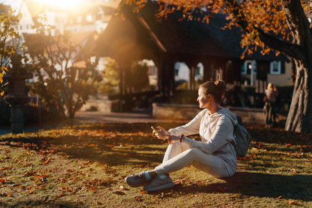 Young Fashionable Teenage Girl With Smartphone In Europian Park In Autumn Sitting At Smiling. Trendy Young Woman In Fall In Park Texting. Retouched, Vibrant Colors. Beautiful Blonde Teenage Girl Wearing Casual Modern Autumn Outfit Sitting In Park In Autumn. Retouched, Vibrant Colors, Brownish Tones.の写真素材