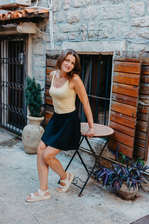 Girl Tourist Walking Through Ancient Narrow Street On A Beautiful Summer Day In MEDITERRANEAN MEDIEVAL CITY, OLD TOWN BUDVA, MONTENEGRO. Young Beautiful Cheerful Woman Walking On Old Street At Tropical Town. Pretty Girl Looking At You And Smilingの写真素材