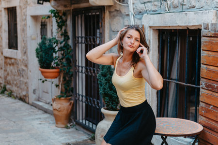 Girl Tourist Walking Through Ancient Narrow Street On A Beautiful Summer Day In MEDITERRANEAN MEDIEVAL CITY, OLD TOWN BUDVA, MONTENEGRO. Young Beautiful Cheerful Woman Walking On Old Street At Tropical Town. Pretty Girl Looking At You And Smilingの写真素材