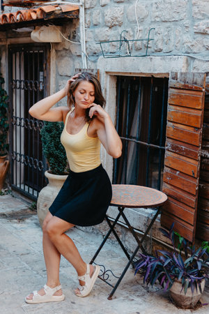 Girl Tourist Walking Through Ancient Narrow Street On A Beautiful Summer Day In MEDITERRANEAN MEDIEVAL CITY, OLD TOWN BUDVA, MONTENEGRO. Young Beautiful Cheerful Woman Walking On Old Street At Tropical Town. Pretty Girl Looking At You And Smilingの写真素材
