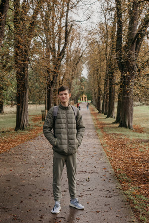 a guy in a jacket stands on an alley in the park during the fall season. Portrait oh handsome teen guy, young man in hoodie, down jacket standing, walking in beautiful golden autumn park, looking at camera. Natural background, colourful leaves, trees. Portrait of young smiling man in casual jacket looks at camera on autumn nature background in countryside or in park. Concept of style, walking in fresh air and unity with natureの写真素材