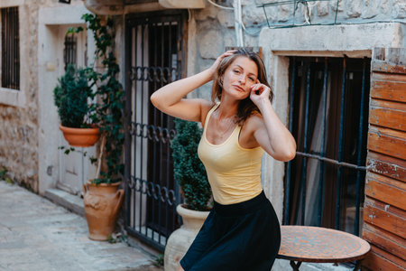 Girl Tourist Walking Through Ancient Narrow Street On A Beautiful Summer Day In MEDITERRANEAN MEDIEVAL CITY, OLD TOWN BUDVA, MONTENEGRO. Young Beautiful Cheerful Woman Walking On Old Street At Tropical Town. Pretty Girl Looking At You And Smilingの写真素材