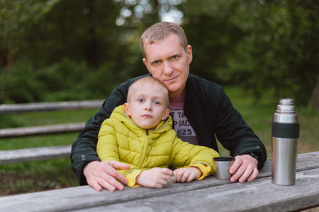 Happy Family: Father And Child Boy Son Playing And Laughing In Autumn Park, Sitting On Wooden Bench And Table. Father And Little Kid Having Fun Outdoors, Playing Together. Father And Son Sitting On A Bench And Talking. Thermosrの写真素材