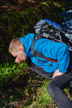 Hiker drinking stream water in mountain. Adult man drinking water from the mountain creek. Tourist wearing casual clothes and sunglasses making a sip of mountain river water from the palms of his hands when hiking.の写真素材