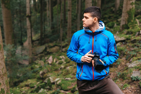 Young Caucasian Male Photographer Taking Picture in the autumn park. Photographer man holding camera in the woods. Photographer man taking a photo outdoor traveling. A photographer with his camera in hand in the mountain forest. The landscape with forests, mountains. Travel or adventure concept.の写真素材