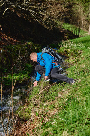 Hiker drinking stream water in mountain. Adult man drinking water from the mountain creek. Tourist wearing casual clothes and sunglasses making a sip of mountain river water from the palms of his hands when hiking.の写真素材