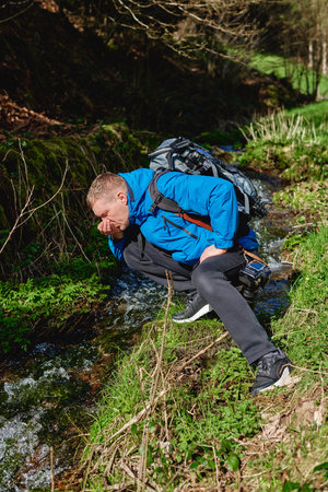 Hiker drinking stream water in mountain. Adult man drinking water from the mountain creek. Tourist wearing casual clothes and sunglasses making a sip of mountain river water from the palms of his hands when hiking.の写真素材