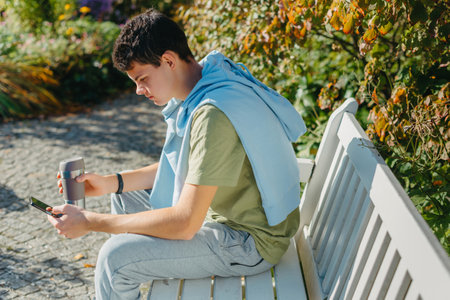 A Teenager Sits On A Bench In The Autumn Park Drinks Coffee From A Thermo Mug And Looks Into A Phone. Portrait Of Handsome Cheerful Guy Sitting On Bench Fresh Air Using Device Browsing Media Smm Drinking Latte Urban Outside Outdoorの写真素材