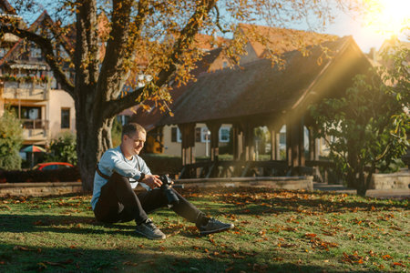 Professional Photographer Taking Picture Of Beautiful Autumn Park. Man Professional Photographer Sit With Camera And With Smartphone In Autumn Park. Retouched, Vibrant Colors, Brownish Tones.の写真素材