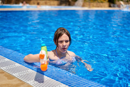 Young pretty caucasian girl with blue eyes shoots with a water gun in the pool. Pretty girl playing...
