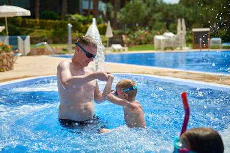 Dad with children splashing in the pool. Father and sons in swimming pool on summer vacation. Child with dad playing in pool. Dad and kid in pool at summer day. Leisure and swimming at holidays. Father have fun with the son in the pool.の写真素材