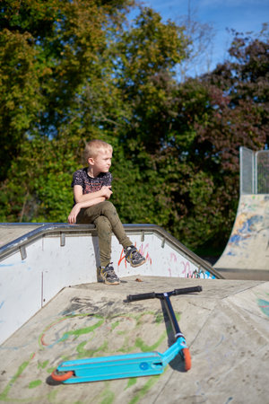 Boy on scooter makes a trick and enjoying his riding in the skate park at cloudy spring day. Young man doing trick on the kick scooter in the park. Full body little boy on the kick scooter on sunny autumn day in skate park. From below of active teen boy with kick scooter standing on ramp in skate park while preparing for performing trick in sunny spring day.の写真素材