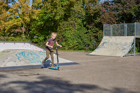 Boy on scooter makes a trick and enjoying his riding in the skate park at cloudy spring day. Young man doing trick on the kick scooter in the park. Full body little boy on the kick scooter on sunny autumn day in skate park. From below of active teen boy with kick scooter standing on ramp in skate park while preparing for performing trick in sunny spring day.の写真素材