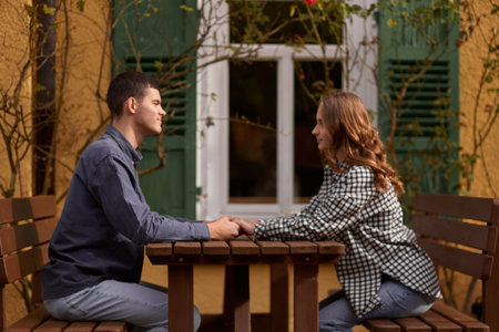 Happy smiling young dating couple having coffee together and enjoying life sitting at table holding hands in street cafe on summer day. Pretty man and woman spending time together enjoying weekend. Dating, young fashion elegant stylish couple sitting at a table in a cafe , gently hold hands, on streets of old european city. American beautiful man hugging caucasian woman and sitting at cafe, hat on table. Concept of happy international couple and dating.の写真素材