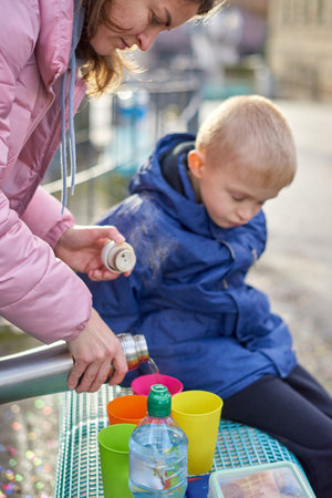 Family Picnic Delight: Cheerful 8-Year-Old Blond Boy in Blue Winter Jacket Sits on Bench While Mom Pours Tea from Thermos, Autumn or Winter. a joyful 8-year-old blond boy in a blue winter jacket, sitting on a bench while his mom pours tea from a thermos into colorful plastic cups.の写真素材