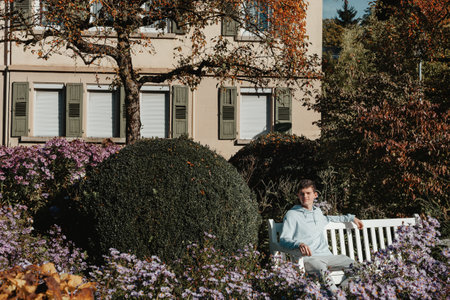 A teenager sits on a bench in the autumn park and looks around. Portrait of cheerful guy sitting on bench in fresh air drinking coffee outside.の写真素材