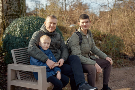 Dad with two sons sitting on a bench in autumn park. Autumnal Family Serenity: Father, 40 Years Old, and Two Sons - Beautiful 8-Year-Old Boy and 17-Year-Old Young Man, Seated in the Parkの写真素材