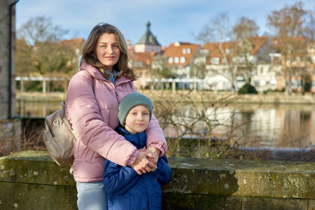 Riverside Family Harmony: Mother, 30 Years Old, and Son - Beautiful 8-Year-Old Boy, Standing by Neckar River and Historic Half-Timbered Town, Bietigheim-Bissingen, Germany, Autumnの写真素材