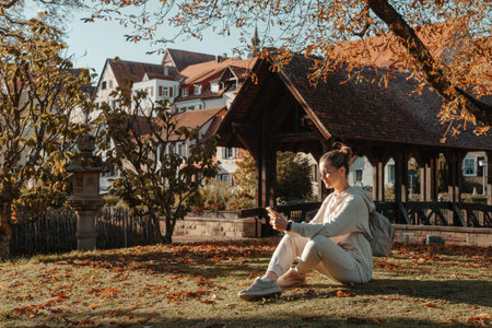 Young Teenage Girl With Smartphone In Park In Autumn Sitting At Smiling. Young Woman In Fall In Park Texting. Retouched, Vibrant Colors. Beautiful Blonde Teenage Girl Wearing Casual Modern Autumn Outfit Sitting In Park In Autumn. Retouched, Vibrant Colors, Brownish Tones.の写真素材