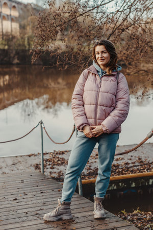 A young woman standing at the shore looking at the river in autumn sunny day. Street view, copy space for text. Happy tourist woman on the bank of the river in autumn in warm clothes. Tourists enjoy their vacation. Romantic look and travel concept. A joyful mood in a girl.の写真素材