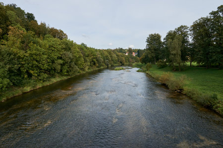 Serene River Landscape with Cozy Cottages Nestled in Lush Greenery Under Dramatic Twilight Sky. Tranquil Riverside Village at Dusk: Picturesque Houses Nestled Among Lush Greeneryの写真素材