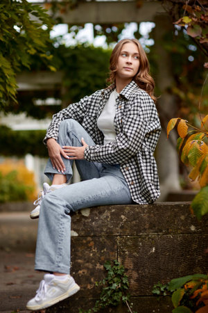 Stylish young woman in casual autumn outfit sitting on a stone wall in a green park with orange...