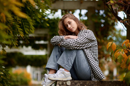Stylish young woman in casual autumn outfit sitting on a stone wall in a green park with orange leaves, wearing a black and white plaid shirt, jeans, and sneakers â outdoor fashion portrait.の写真素材