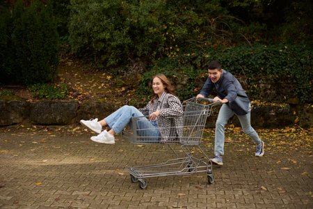 Happy young couple having fun with shopping cart outdoors in autumn park, wearing stylish casual outfits with denim and flannel, laughing and enjoying joyful moment, surrounded by autumn foliage and warm natural lighting.の写真素材