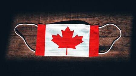 Canada National Flag at medical, surgical, protection mask on black wooden background. Coronavirus Covidâ19, Prevent infection, illness or flu concept photo. State of Emergency, Lockdown...の写真素材