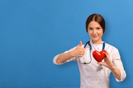 Young doctor girl with white coat uniform and stethoscope holding red heart and showing thumbs up on...