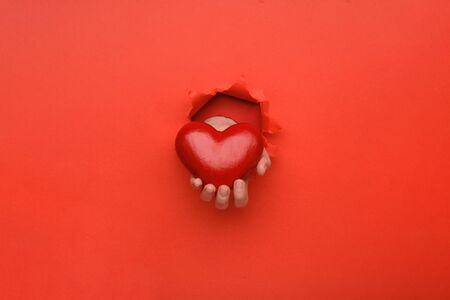 Hand holding red heart on torn red paper wall. Copy space aside for your advertising and offer or sale content. Valentines day, mother day, social advertising conceptの写真素材