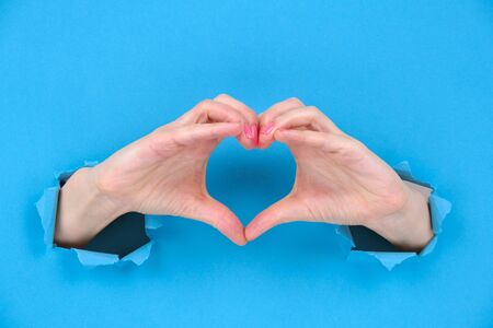 Female hands show heart symbol through ragged holes of a blue torn paper background. Creativity, a...