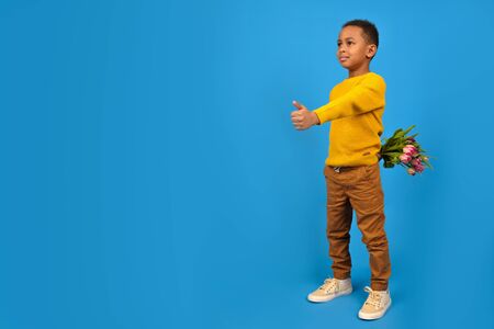 African-American boy with bouquet of tulips hides behind his back, finger-up gesture of approval or like on blue background. concept mothers Day, fathers day, Valentines day gift for womanの写真素材