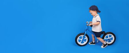 Happy baby smiling on a runner learns to keep balance blue bike (run bike), in white polo and blue shorts on a blue backgroundの写真素材