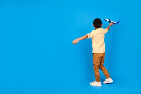 Happy African-American boy plays with a toy plane, puts his hands aside, imagines himself flying in a plane against a blue background. Family travel and tourism, summer holidays conceptの写真素材