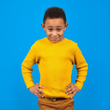 Cheerful happy emotional African-American boy smiles and laughs at camera in yellow sweater on blue Studio background. Concept of human emotions and happiness.の写真素材