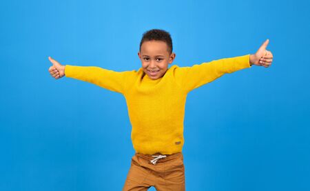 Happy African-American boy rejoices and smiles at victory or achievement, raises his hands up, on a blue studio homogeneous background. concept of winning, achieving the goal of striving to win.の写真素材