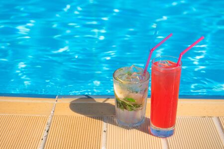 Two refreshing cocktails Mojito and Cosmopolitan stand on the edge of the pool in the summer heat...