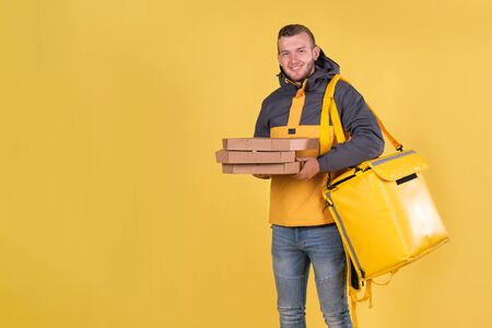 Food delivery smiling caucasian young man in yellow jacket and with thermal bag on his shoulders on yellow background holds an order of three pizzas in craft box for customer. Concept express deliveryの写真素材