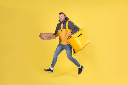 Blond unshaven teen food delivery boy and courier in yellow clothes with thermo bag on his back holds an order for customer and runs to deliver order on time in 15 minutes. Food delivery in cityの写真素材