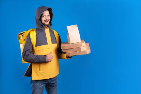 Unshaven dark-haired caucasian courier in yellow windbreaker holds an order of pizza and coffee and refrigerator bag for food, delivery man shows an approving hand gesture symbolizing good delivery.の写真素材