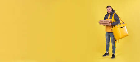 Food delivery smiling Caucasian young man in yellow jacket and with thermos bag on his shoulders on...