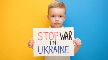 Portrait Blond little boy calls to Stop war in Ukraine, raises banner with inscription stop the war in Ukraine standing on blue-yellow studio background. No war, stop war, russian aggression.の写真素材