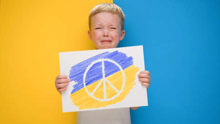 Boy crying on yellow-blue background of flag Ukraine holding banner Pride peace symbol with colors of flag of Ukraine. Child against war, crisis Ukraine, humanitarian catastrophe, Russian aggression.の写真素材