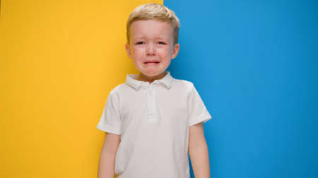 Portrait small blond boy crying on yellow-blue background flag of Ukraine. Crisis in Ukraine, war...