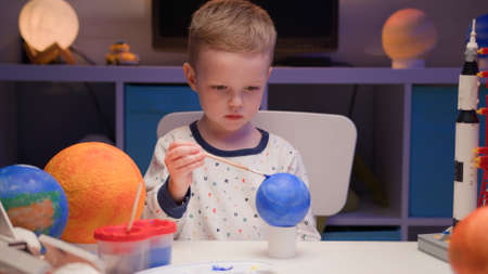 Blond boy paint planet solar system Pluto with colorful paint sitting home table in evening, planet solar system, spaceships and space shuttle from constructor around. Cosmonautics Day on April 12.の写真素材
