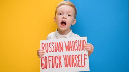 Portrait small blond boy raising banner with inscription shouts slogan Russian warship, go fuck yourself, standing on blue-yellow background of studio. No war, stop war, Russian aggression.の写真素材
