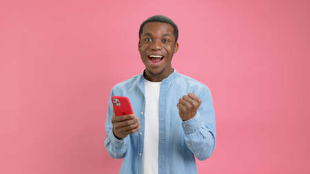 Shocked happy victory young African American 20 years old in denim shirt, emotion of winning casino or sports betting in mobile application poses in isolation on pink background. Emotions of victory.の写真素材