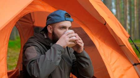 portrait bearded traveler man sitting in an orange tent wearing blue cap and gray jacket in nature...
